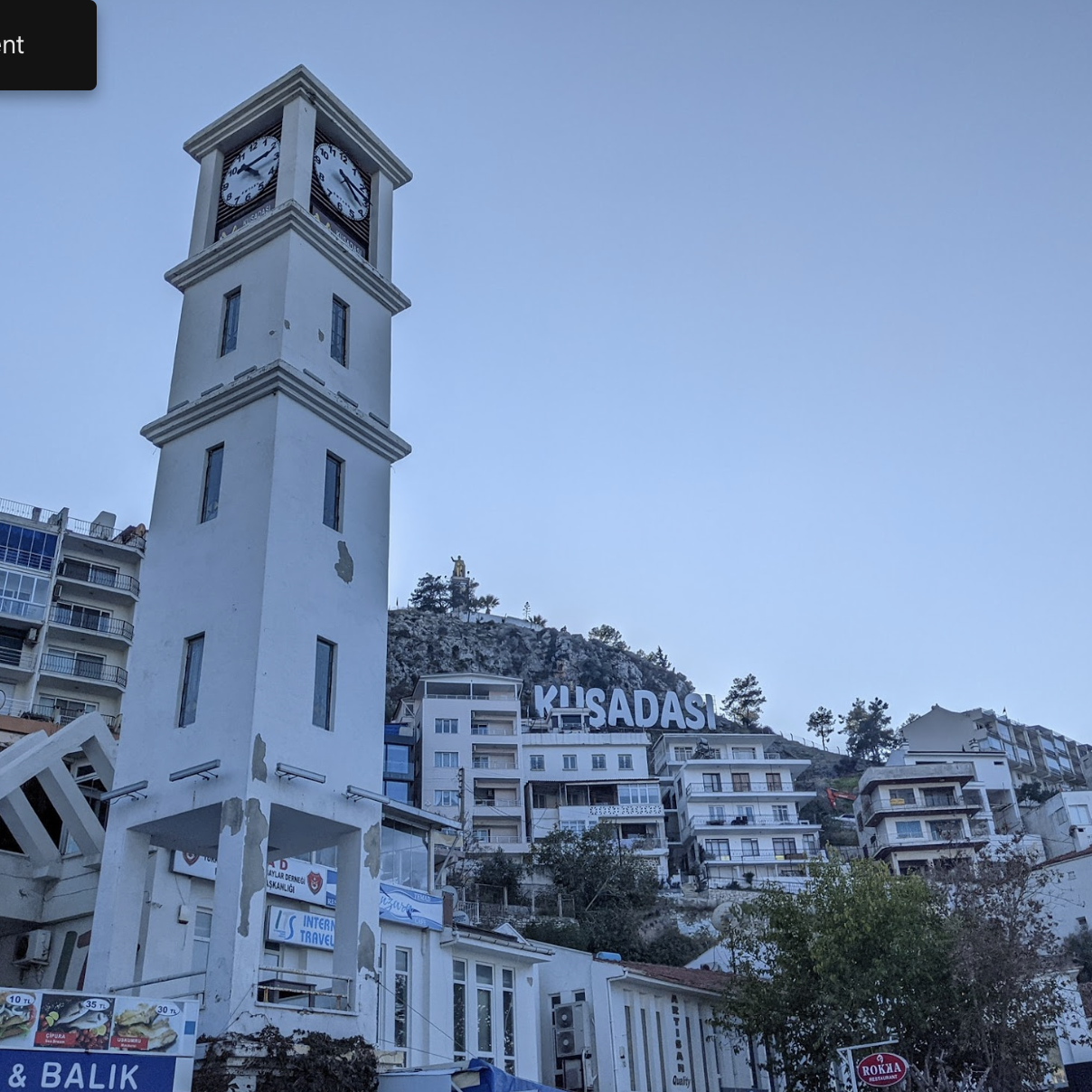 საათის კოშკი კუშადასის ქალაქის ცენტრში გორაკზე შენობებით და კუშადასის ნიშნით, მოწმენდილი ცის ქვეშ.